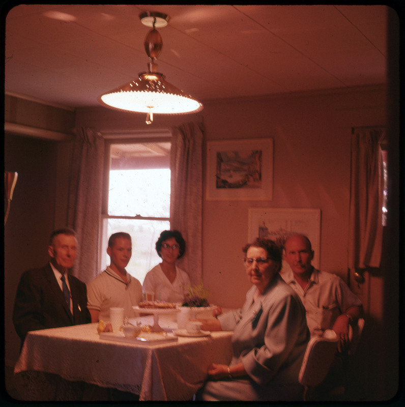 A photographic slide of five people sitting at a table celebrating. There is a cake with candles. Donald Crabtree is sitting at the table and the photo is slightly blurry.