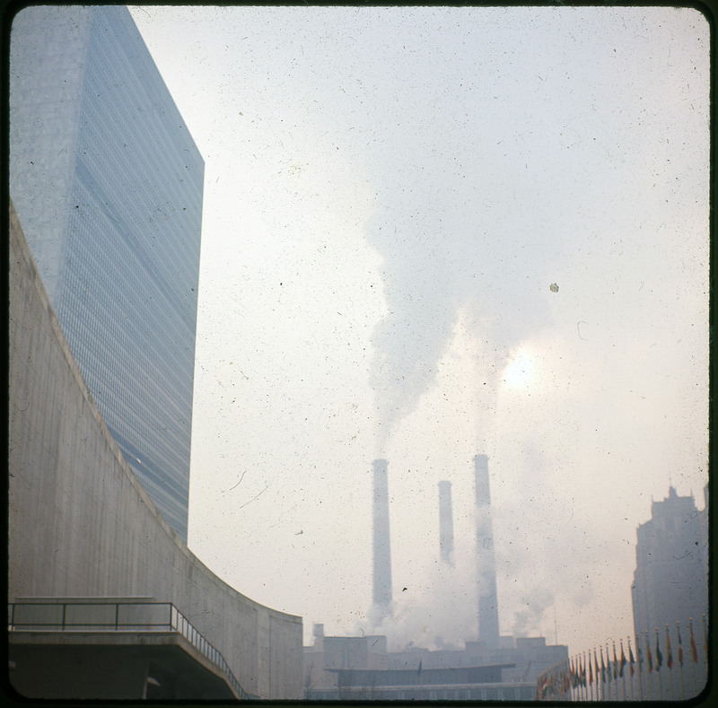 A photographic slide of a factory and the smoke its producing. There is a large building on the side of the photo.