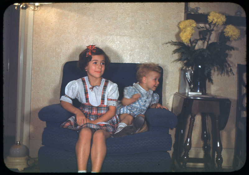 A photographic slide of two children sitting together on a chair. There is a vase of flowers next to them.