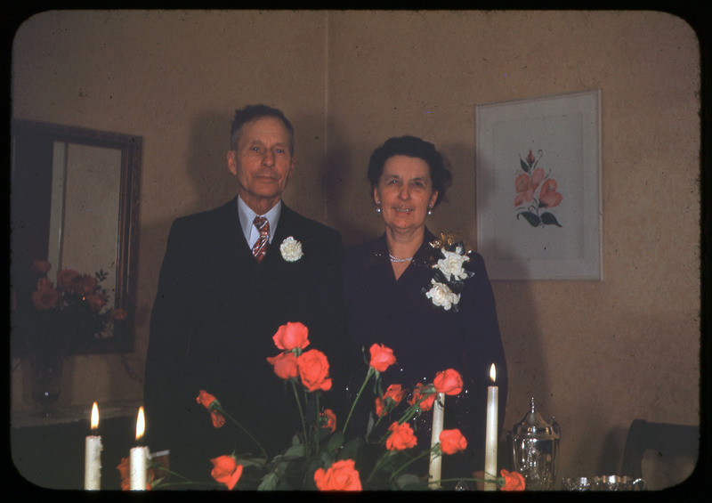 A photographic slide of a couple posing together with corsages. There is a vase of flowers on the table in front of them.