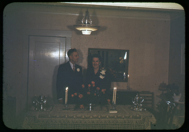 A photographic slide of a couple standing in front of a table. The table is set with candles, tea sets, and flowers. The couple is wearing corsages.