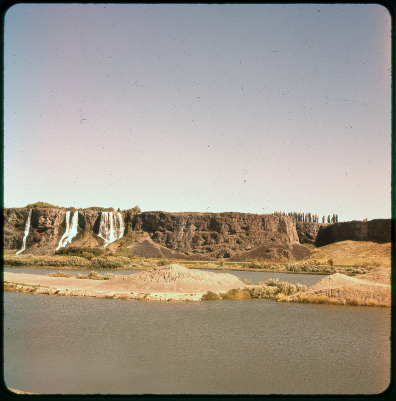 A photographic slide of a river canyon. There are three waterfalls on the rock and a sand dune in the center.