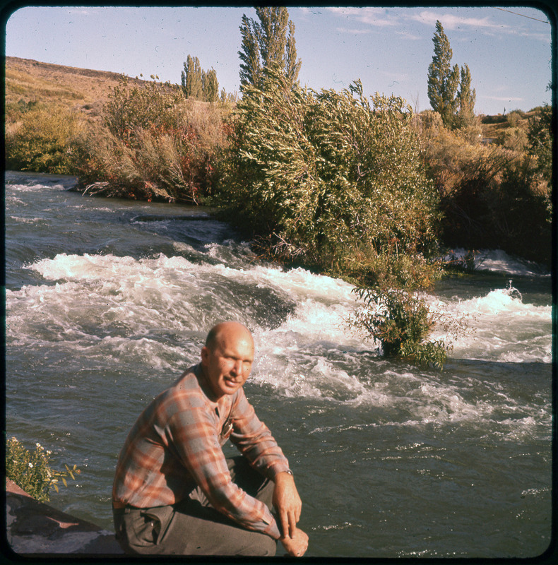 A photographic slide of Donald Crabtree in front of a raging river. There are many trees and bushes along the river's edge.