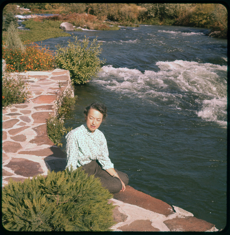 A photographic slide of Evelyn Crabtree sitting on a stone edge next to a river. The river is flowing through rapids in the background.