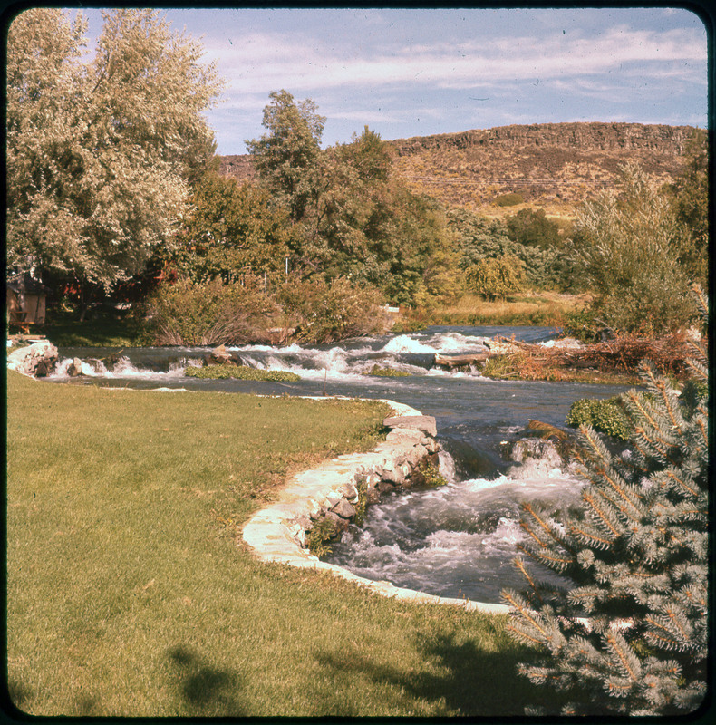 A photographic slide of a rushing river with a lawn next to it. There are many trees and bushes along the edge. There is a rocky mountain in the background.