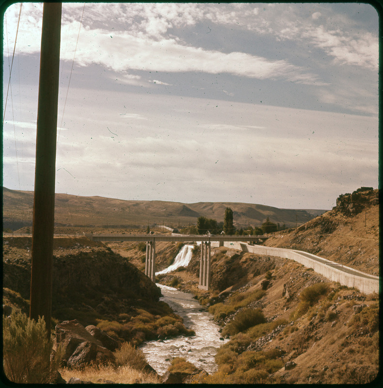 A photographic slide of a large dam and waterfall. There are many mountains in the background and an aqueduct.