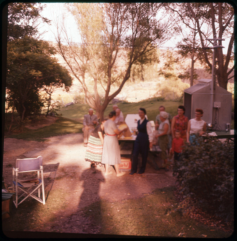 A photographic slide of a group of people surrounding a picnic table. There is a large lawn and trees in the background. People are standing and talking.