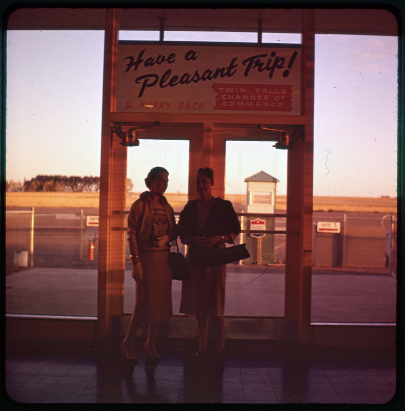 A photographic slide of Evelyn Crabtree and another woman standing together in front of an airport doorway. There are gates in the background for the airport.