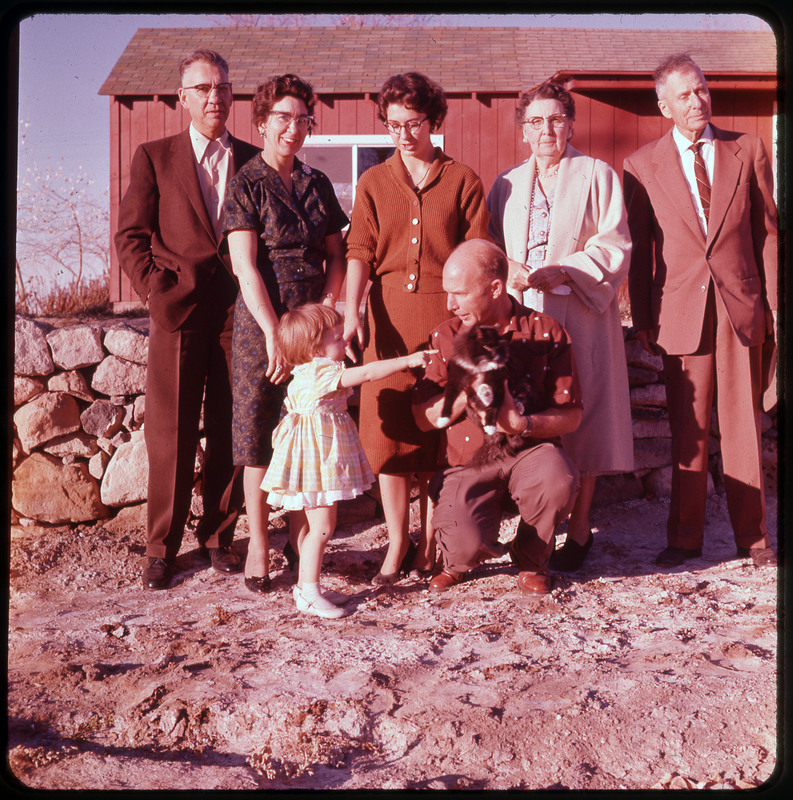 A photographic slide of Donald Crabtree holding a cat and a child pointing to it. They are crouched down and there are others in the photo with their heads not pictured.