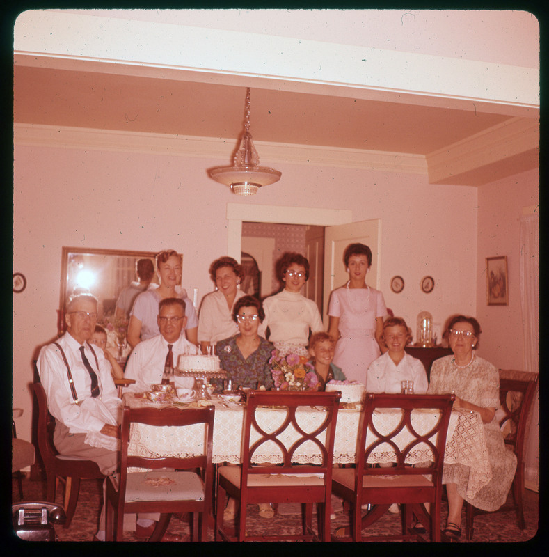 A photographic slide of ten people sitting and standing around a table. They are dressed nicely and there are cakes with candles on the table.