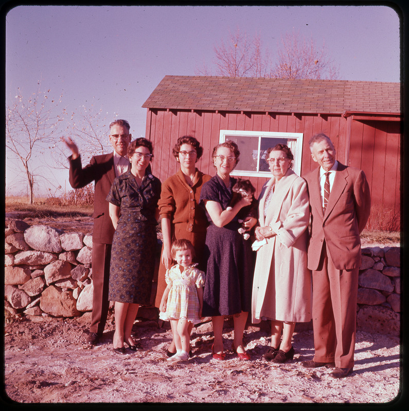 A photographic slide of a family standing outside a red house together. Evelyn Crabtree is holding a cat and there are seven people pictured.