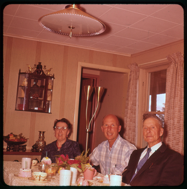 A photographic slide of Donald Crabtree smiling and two others sitting with him at a table.