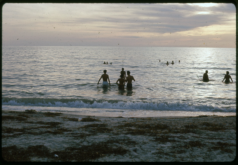 A photographic slide of a beach with people swimming in the ocean at sunset.