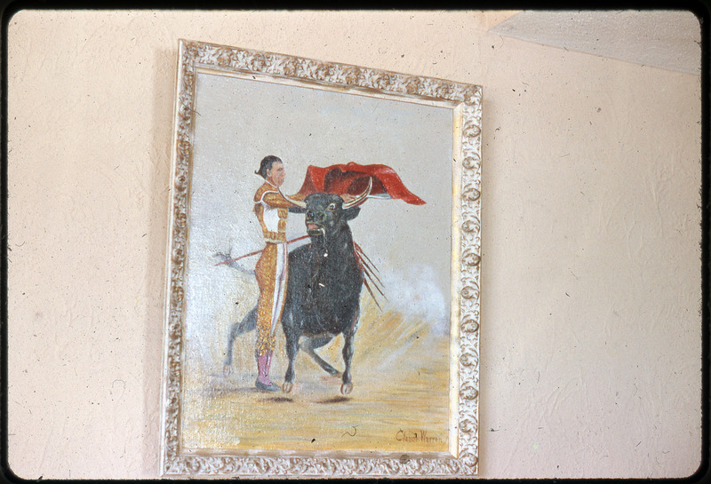 A photographic slide of a bull and a matador man holding a red cape over the bull. The painting is on the wall.