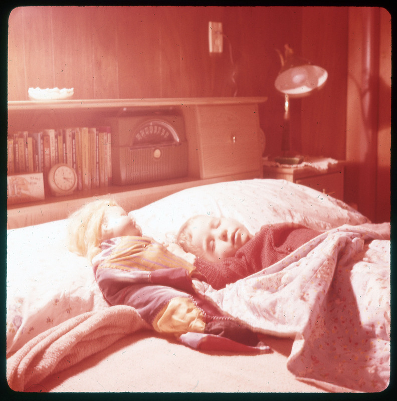 A photographic slide of a child sleeping in bed with a doll next to them. The doll is also laying in bed as if sleeping.