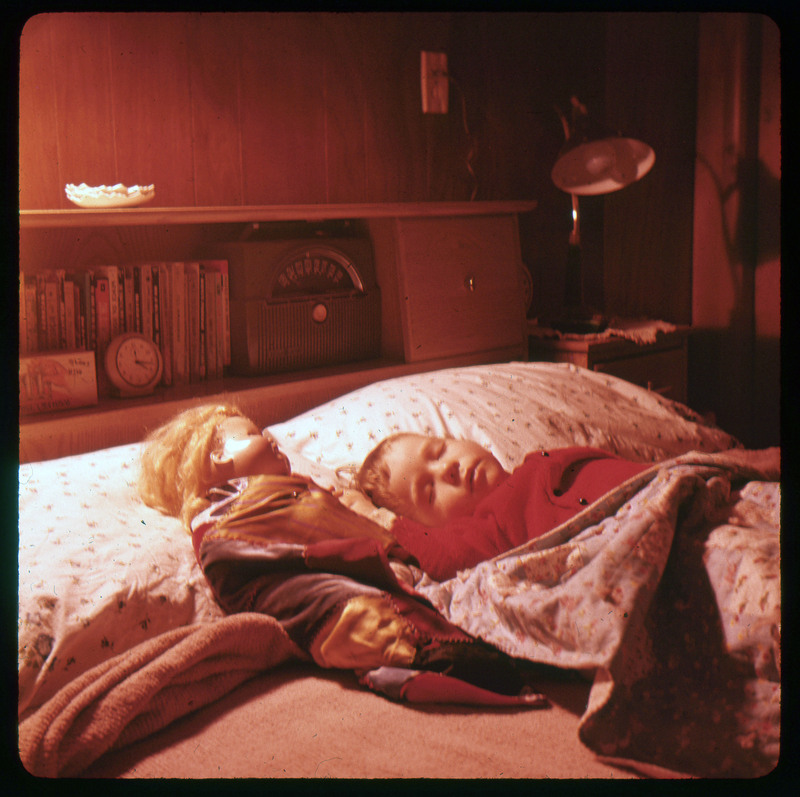 A photographic slide of a young child laying in bed sleeping next to a doll.