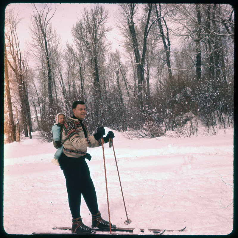 A photographic slide of a person skiing with a child in a backpack on his back. It is a snowy scene.