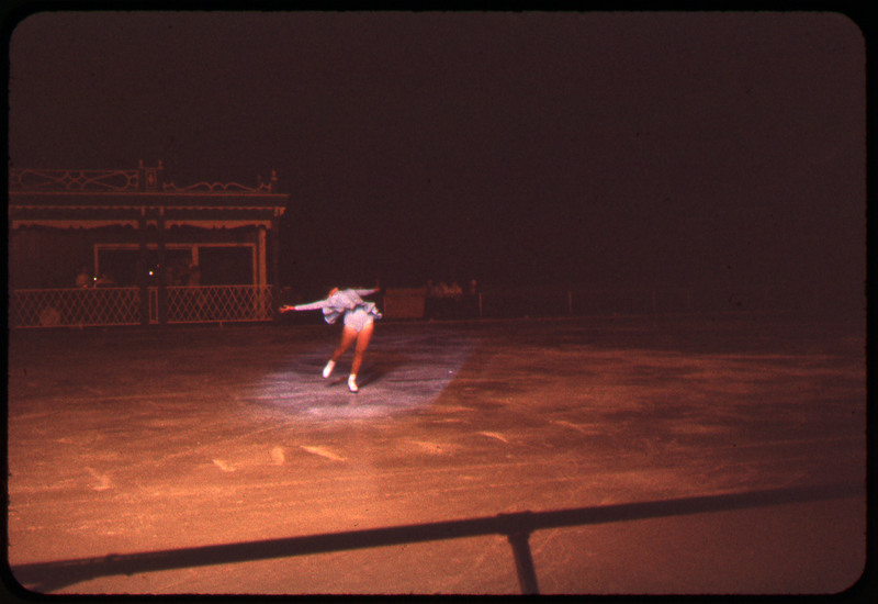 A photographic slide of a woman doing a trick while figure skating. There is a structure behind her.