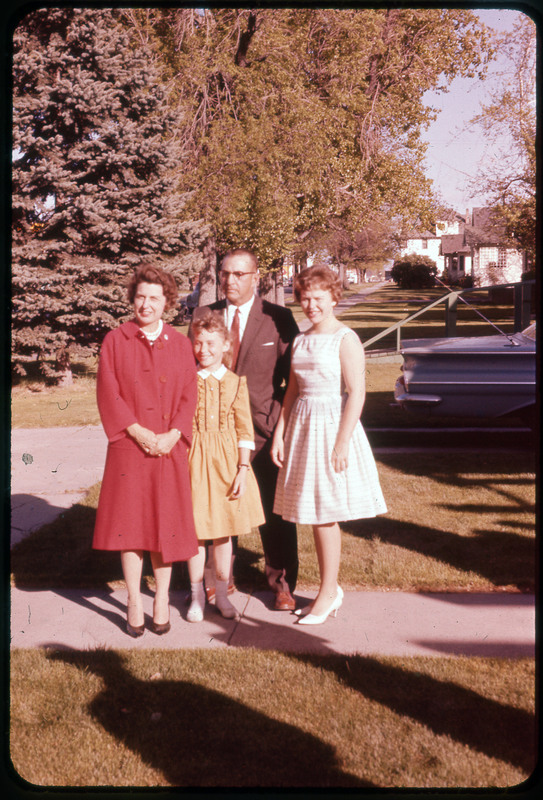 A photographic slide of a family standing together outside. They are standing on a sidewalk with houses in the background and a car parked nearby.