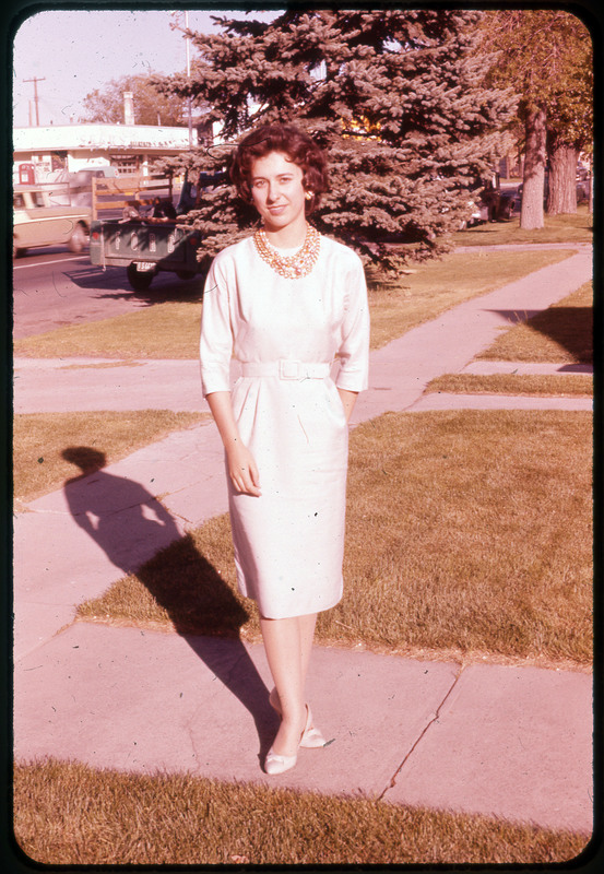 A photographic slide of a woman standing on the sidewalk outside a house. She is wearing a white dress.