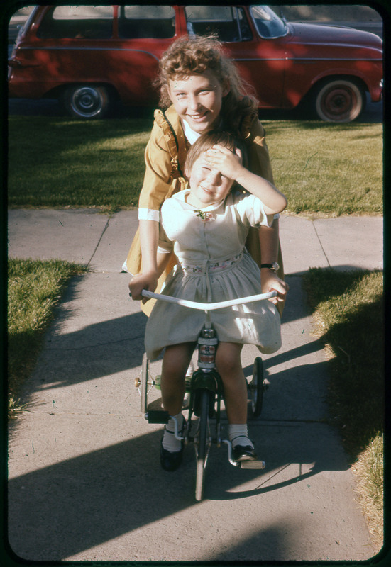 A photographic slide of two children playing together outside on an tricycle. They are smiling.