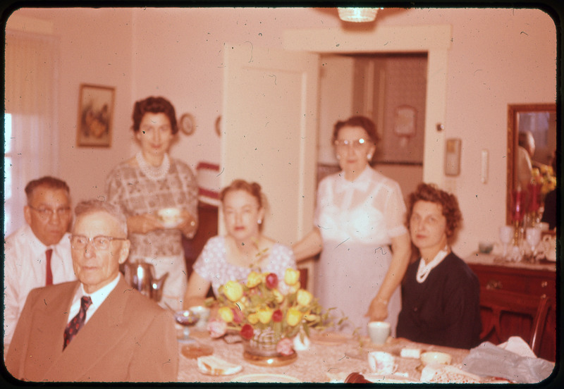 A photographic slide of a group of people drinking tea or coffee together at a table. There are flowers on the table and they are dressed nice.