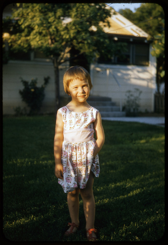 A photographic slide of a child standing outside on a lawn in front of a house.