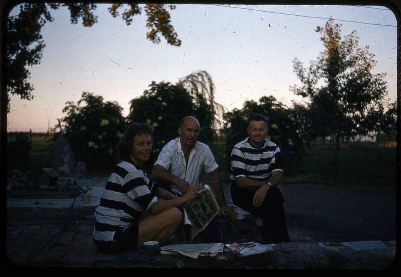 A photographic slide of three people sitting together outside reading magazines and the newspaper. Donald Crabtree is pictured and two others wearing matching striped shirts.