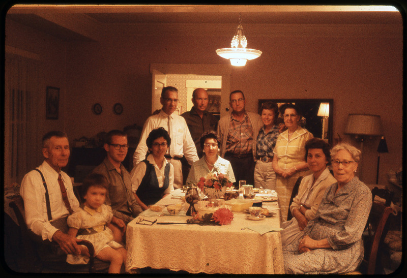 A photographic slide of people standing and sitting around a nice dinner table. There are flowers on the table and a lace tablecloth.