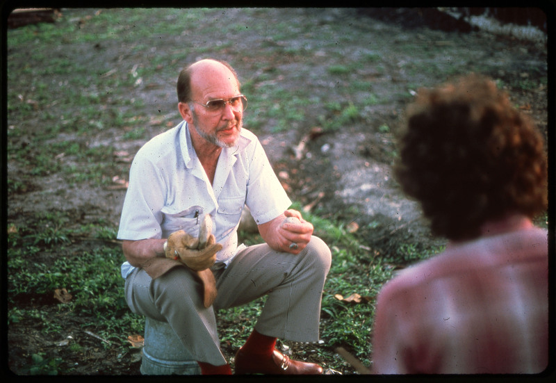 A photographic slide of Donald Crabtree talking while giving a flintknapping demonstration. He is sitting on a bucket and holding a hammerstone.