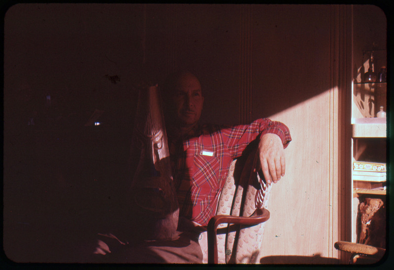 A photographic slide of Donald Crabtree sitting on a chair with a wooden carved sculpture. The carving is of a face.