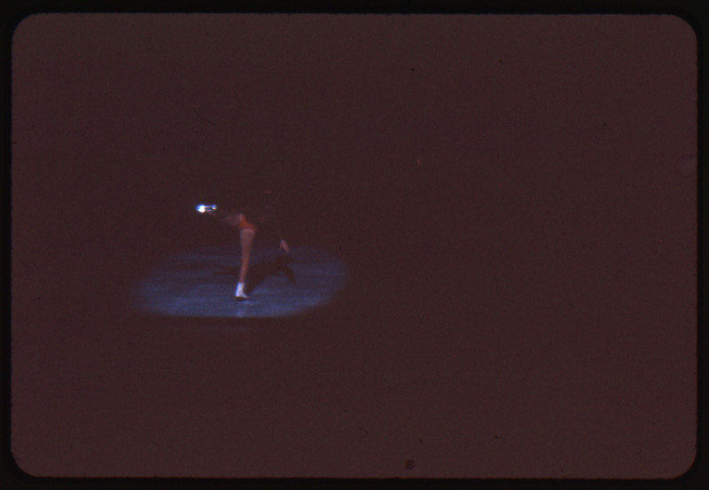 A photographic slide of a person doing a trick while figure skating. There is a spotlight on them.