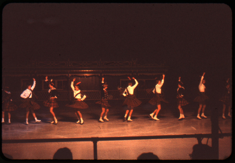 A photographic slide of a group of figure skaters in a line doing a dance. They are wearing matching outfits.