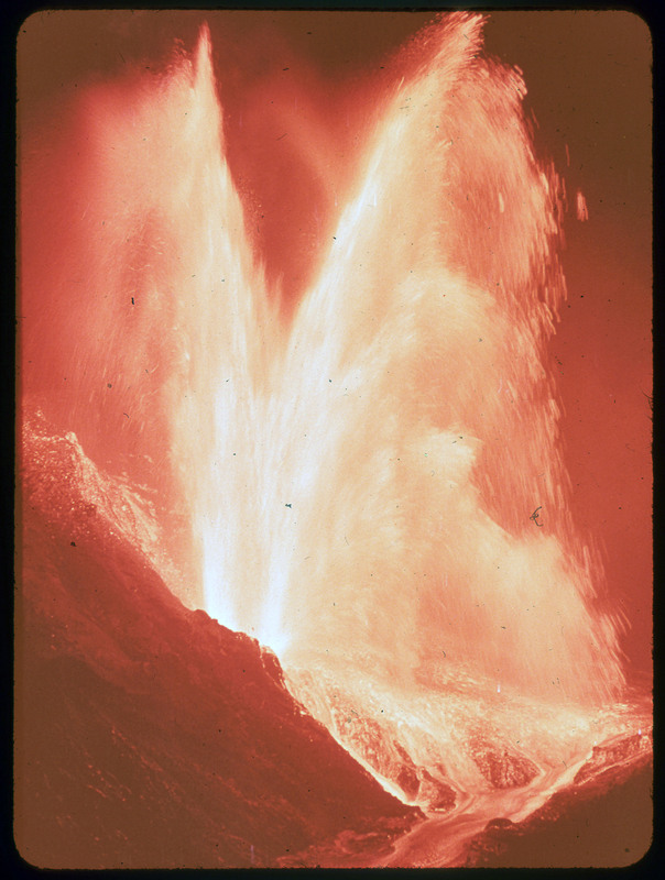 A photographic slide of lava erupting from a volcano. It is bright orange and there is lava flowing down in a stream.