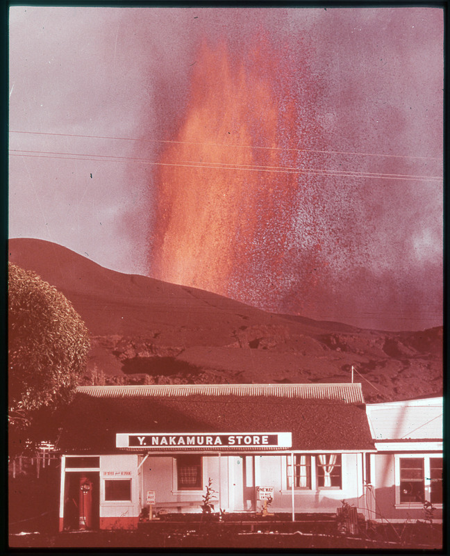 A photographic slide of a large lava eruption behind a mountain. There is a store in front.