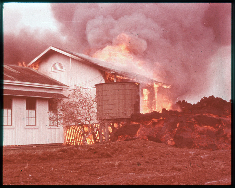 A photographic slide of a lava flow hitting a house and catching it on fire.