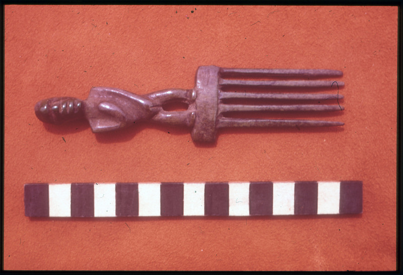 A photographic slide of a comb artifact on a red surface. The comb has a carving of a face.