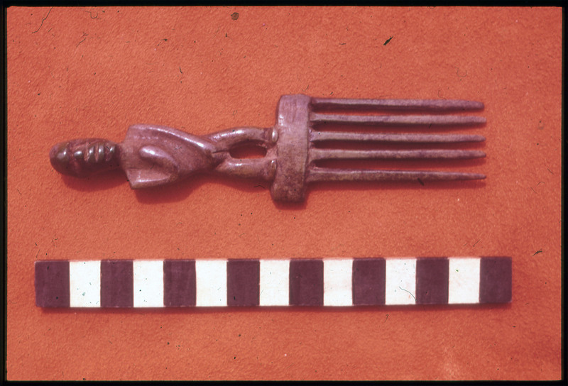 A photographic slide of a comb artifact on a red surface. The comb has a carving of a face.