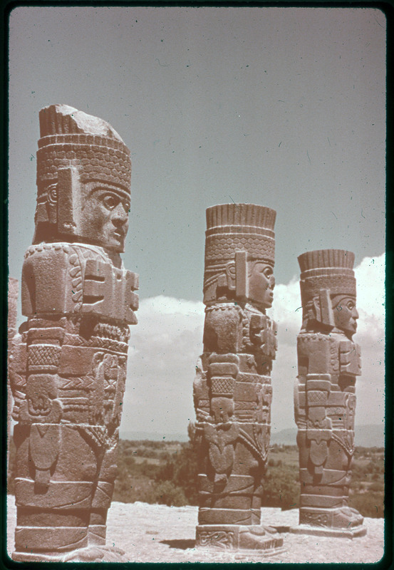 A photographic slide of three stone mesoamerican statues of people. They are on top of the mountain.