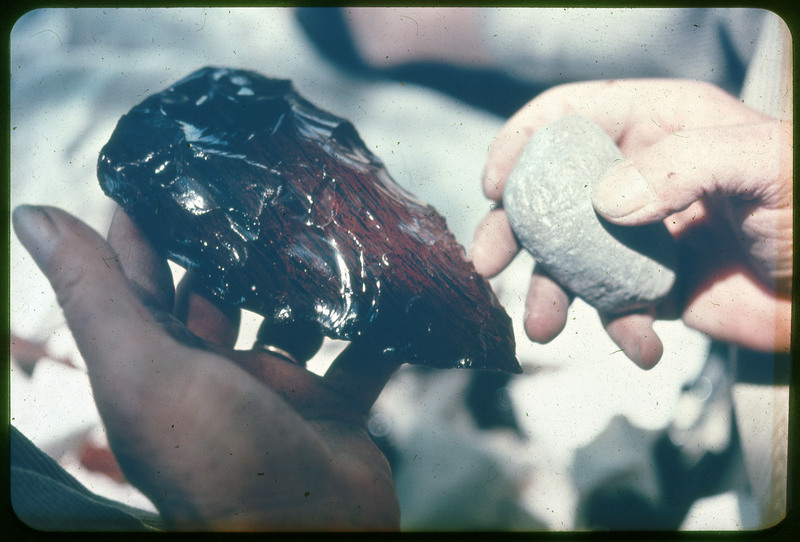 A photographic slide of a person holding a red, large biface and a white, small hammerstone.