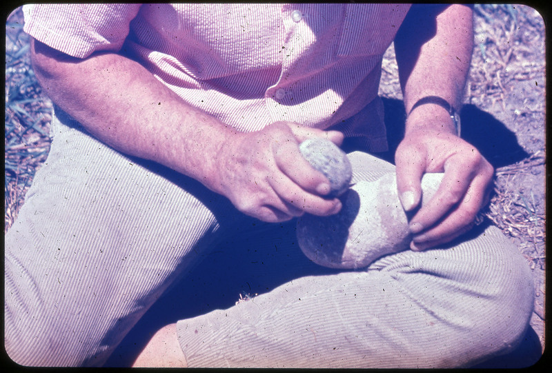 A photographic slide of a person using two large stones to flintknap. He is sitting outside.