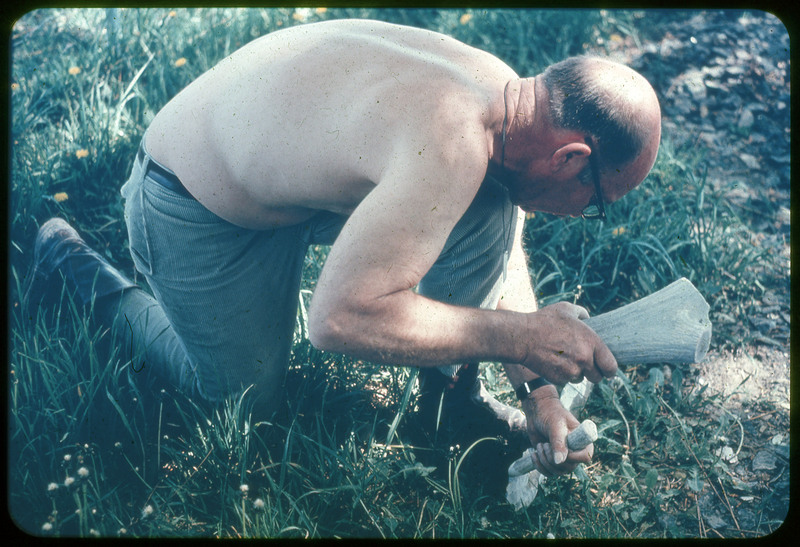 A photographic slide of Donald Crabtree flintknapping a very large biface using a large antler. He is kneeling in grass.