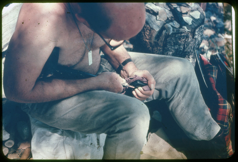 A photographic slide of Donald Crabtree flintknapping using a large copper tipped shoulder crutch. There is a log with flakes on it and a coat by his feet.