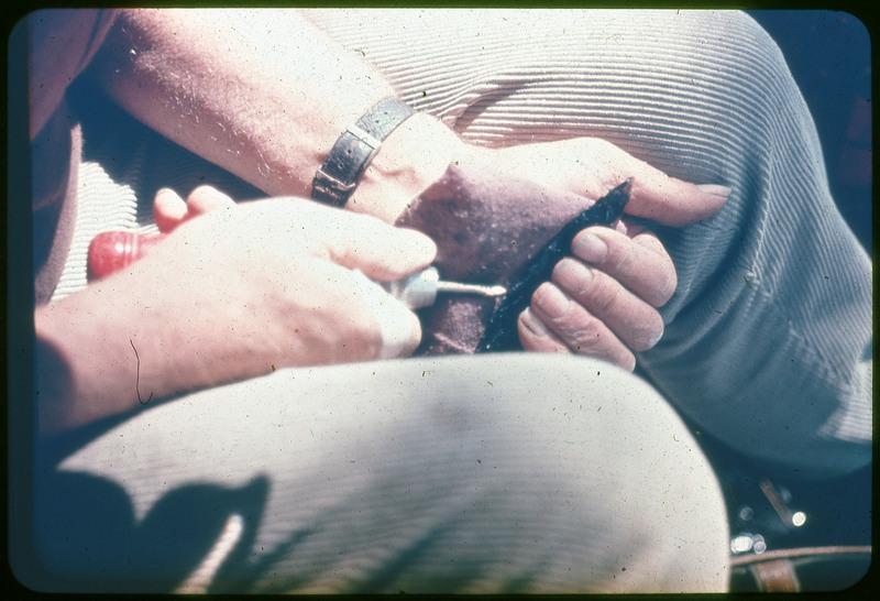 A photographic slide of Donald Crabtree flintknapping a small black biface. He is using a copper tipped tool to flake the edges.