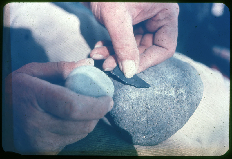 A photographic slide of a person holding a very small biface in the process of flintknapping. The person also holds a hammerstone.