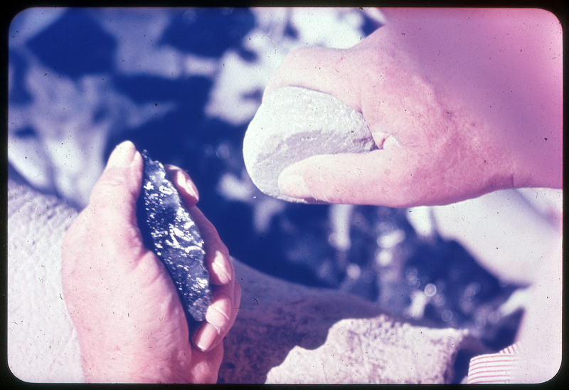 A photographic slide of a person using a hammerstone to flintknap a black obsidian biface over a leather lap pad.