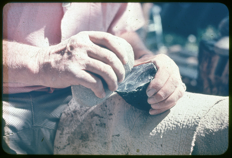 A photographic slide of a person flintknapping a large piece of obsidian. The person is using a hammerstone to remove flakes.