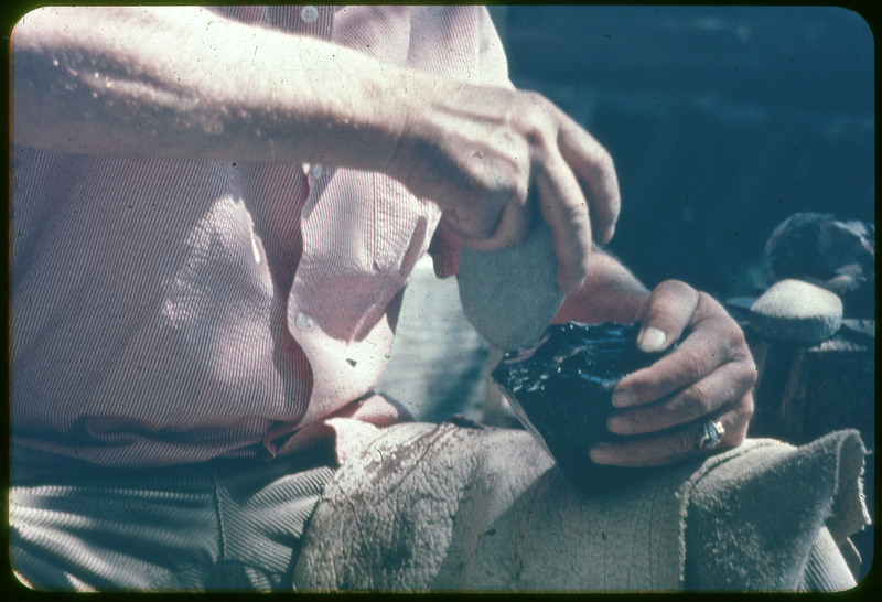 A photographic slide of a person flintknapping a large black piece of obsidian. The person is striking with a hammerstone from above.