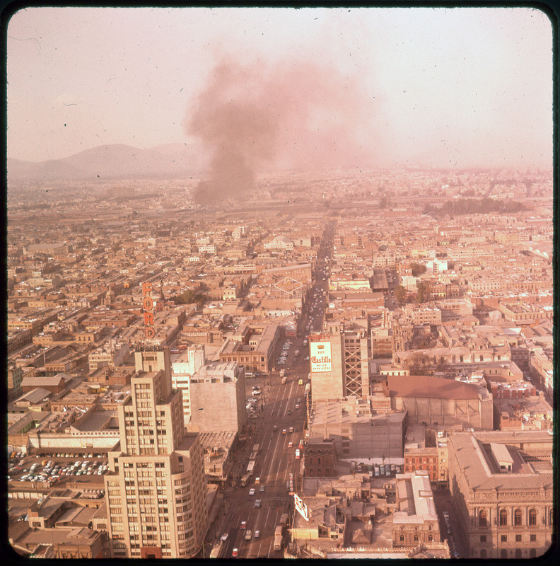 A photographic slide of an industrial-like city. There are many buildings, smoke in the background, and a busy street.