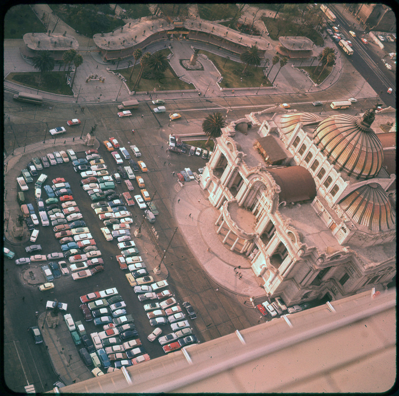 A photographic slide of a full parking lot of cars. There is a large building with columns and ornate features. There is a road with cars on it.